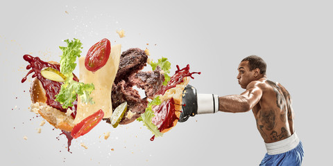 Fight the fast food. Burger's crashing by the boxer isolated on white background. Combination of buns, vegetables, sauce and cutlet is broken by sportsman. Healthy eating, diet and sport concept.