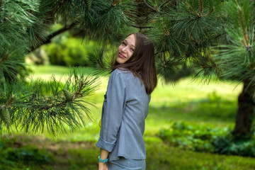 The young woman in gray business suit, in trousers among big green pines in the park. Fashion and style. Portrait.