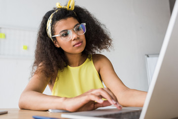 Wall Mural - selective focus of attractive african american woman in glasses using laptop