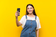© luismolinero - Young woman in dungarees over isolated yellow background making a selfie