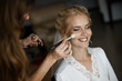 A bride is laughing while a makeup artist is putting a makeup on her face.