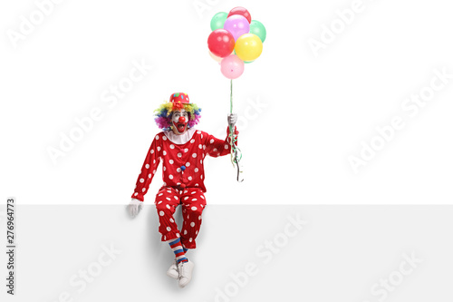 Fotomural Cheerful clown sitting on a white banner and holding a bunch of balloons