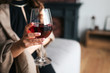 © estradaanton - Cut view of woman's hand holding glass of red wine. Model wear black dress and brown shawl. Woman in living room alone.