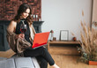 © estradaanton - Young businesswoman work at home. Sit and hold laptop on knees. Galss of red wine in right hand. Busy attractive woman remote work. Typing on keyboard.