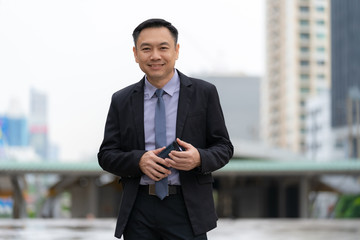 Wall Mural - Asian Businessman standing and holding mobile phone with business office buildings in the city background