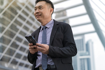 Wall Mural - Asian Businessman standing and holding mobile phone with business office buildings in the city background