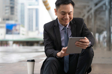 Wall Mural - Asian Businessman sitting and holding digital tablet with business office buildings in the city background