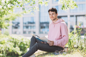 Young handsome student man reading a book in a city park or university campus. Education and modern lifestyle concept