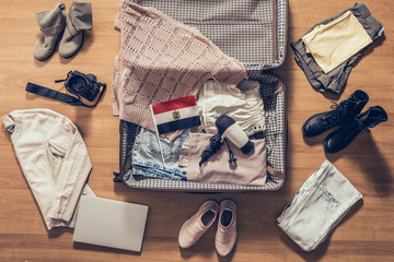 Woman's clothes, laptop, camera and flag of Egypt lying on the parquet floor near and in the open suitcase. Travel concept