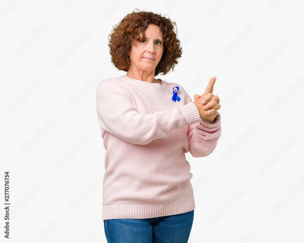 Middle ager senior woman wearing changeable blue color ribbon awareness over isolated background Holding symbolic gun with hand gesture, playing killing shooting weapons, angry face