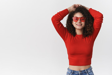 Wall Mural - Stylish carefree charismatic european woman dark curly hairstyle hold hands behind head relaxed joyfully smiling toothy wear cropped top red sunglasses get ready weekend party, white background