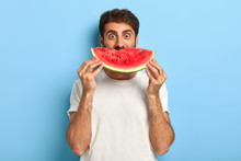 Funny Man Holds Half Of Red Watermelon Near Face, Has Glad Surprised Look, Dressed Casually, Has Healthy Summer Nutrition. European Guy Poses With Tasty Tropical Fruit Against Blue Background