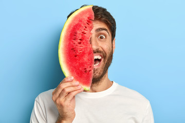 Wall Mural - Isolated shot of smiling young male has natural summer snack, holds slice of fresh watermelon near face, has perfect mood, wears white t shirt, isolated on blue. Seasonal food, organic nutrition