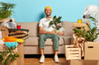 © wayhome.studio  - Satisfied guy poses in empty room on sofa, holds houseplant wrapped in polythene, being happy home owner, busy on moving day with unpacking, rents flat, poses against blue wall. Ownership concept