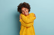 Lovely happy African American woman daydreams, embraces herself, recalls romantic date, feels comfort, keeps hands across body sensually, has broad smile, wears yellow sweater, poses indoor.