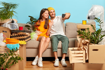 Relocating and changing house concept. Happy newlywed couple move to new abode, pose on cozy couch, glad live together, make selfie portrait on modern smart phone, empty room with packages around.