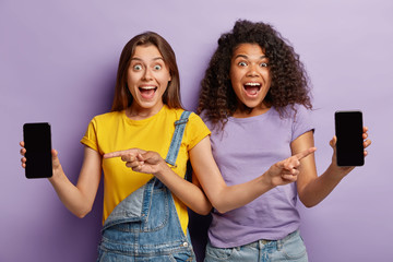 Joyful young multiethnic women point at smartphone display screens, demonstrate with excitement last digital trends, advice cool application, stand next to each other, wears stylish clothing.