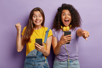 Triumphing joyful female friends clench fists, point at camera, being advanced users of modern technlogies, dressed in stylish outfit, isolated on purple background. Good mood, leisure concept