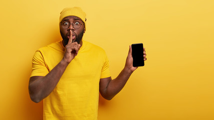 Indoor shot of surprised dark skinned young man makes silence gesture, tells secret, shows modern gadget with blank screen for your promotion or advertising text, isolated on yellow background.