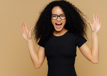 Half Length Shot Of Pleased Overjoyed Woman Dances And Moves Positively, Raises Palms, Exclaims With Happiness, Wears Black T Shirt, Spectacles, Isolated On Beige Wall. Excited Female Models Moves