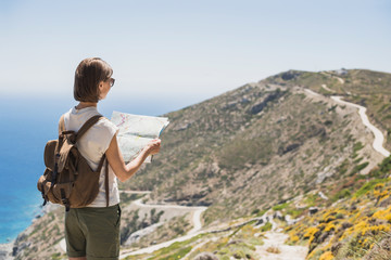 Wall Mural - Woman traveler with backpack holding map. Travel, tourism, summer holidays, active lifestyle concept. Hipster tourist girl looking for hiking route with sea and mountain at background