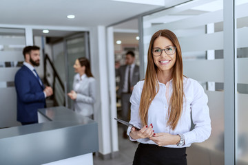 Charming Caucasian businesswoman dressed in shirt and skirt standing on hallway and holding tablet. In background her colleagues standing and chatting.