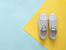 White Leather Sneakers On Blue And Yellow Background. Pair Of Fashion Trendy White Sport Shoes Or Sneakers With Copy Space For Text On Colorful Background. Overhead Shot Of New White Sneakers.Flat Lay