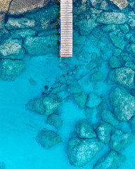 An aerial view of the beautiful Mediterranean Sea, with a wooden pier and a rocky shore, where you can see the textured underwater corals and the clean turquoise water of Protaras, Cyprus