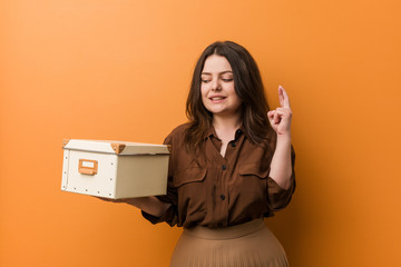 Wall Mural - Young curvy plus size woman holding a box crossing fingers for having luck