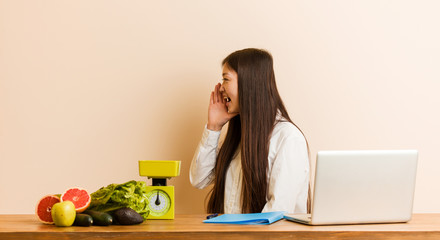 Wall Mural - Young nutritionist chinese woman working with her laptop shouting and holding palm near opened mouth.