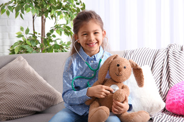 Poster - Cute child imagining herself doctor while playing with stethoscope on sofa in living room