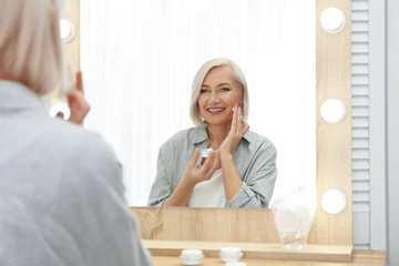 Portrait of charming mature woman with healthy beautiful face skin and natural makeup applying cream indoors