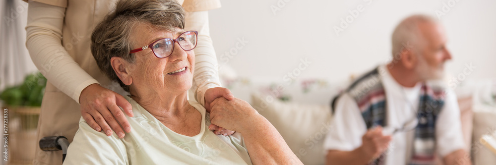 Panoramic view of elderly lady on wheelchair holding hands with supporting volunteer standing behind her - obrazy, fototapety, plakaty 