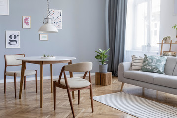 Design scandinavian home interior of open space with stylish chairs, family table wooden commode, gray sofa, accessories and mock up posters gallery wall. Gray background walls. Retro cozy home decor.