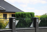 Fototapeta Kamienie - Murowane ogrodzenie z cegły i stali, dach budynku i zielone drzewa.