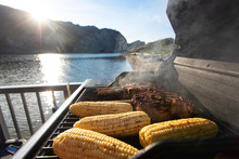 Steak And Corn On The Cob Cooking On An Outdoor Barbecue As The Sun Sets Over A Lake