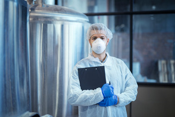 Portrait of technologist in white uniform with hairnet and protective mask and gloves standing in pharmaceutical or food factory with arms crossed.