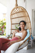 © Liubov Levytska - Cheerful tender happy woman dark curly hair enjoy vacation booked cozy house patio summer vacation lying comfortable rattan lounge hanging wicker chair smiling delighted rest contemplate garden