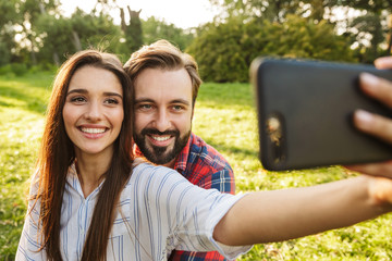 Poster - Image of happy couple man and woman taking selfie photo on cellphone while resting in green park