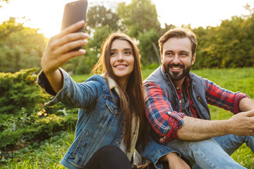 Poster - Image of cute couple man and woman taking selfie photo on cellphone while resting in green park