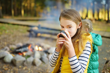 Wall Mural - cute young girl drinking tea and roasting marshmallows on stick at bonfire. child having fun at camp