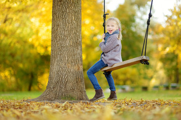 Wall Mural - Cute young girl having fun on a swing in sunny autumn park. Family weekend in a city.