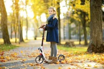 Wall Mural - Adorable young girl riding her scooter in a city park on sunny autumn evening. Pretty preteen child riding a roller.