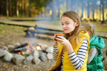 Wall Mural - cute young girl drinking tea and roasting marshmallows on stick at bonfire. child having fun at camp