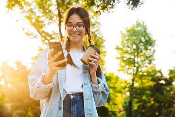 Canvas Print - Happy cheery smiling young teenage girl student walking outdoors in beautiful green park drinking coffee using mobile phone.