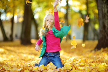 Wall Mural - Adorable young girl having fun on beautiful autumn day. Happy child playing in autumn park. Kid gathering yellow fall foliage.