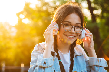 Poster - Pleased happy young teenage girl student sitting outdoors in beautiful green park listening music with headphones.