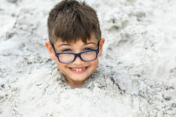 A little boy buried in the sand at the beach. He is cute and wearing eyeglasses