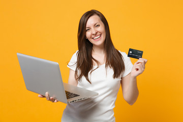 Wall Mural - Portrait of laughing young woman in white casual clothes holding laptop pc computer, credit bank card isolated on yellow orange wall background in studio. People lifestyle concept. Mock up copy space.