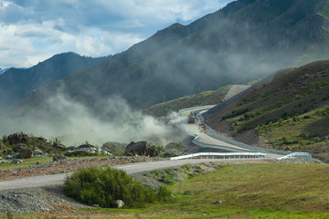 Landscape in the mountains of Altai, disturbed by two large trucks filled with rubble, rising up for the hill, a large amount of dirty dust flies and during construction of road in hard-to-reach place